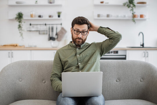 Confused bearded male wearing eyeglasses and casual attire shrugging shoulders while sitting on couch with modern laptop and looking at camera. Concept of doubt, people and technology.