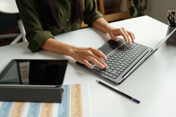 Hands of teacher typing on laptop keyboard while sitting by desk with tablet and document with...