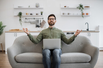 Caucasian man meditating with closed eyes while working on portable laptop at living room. Bearded...