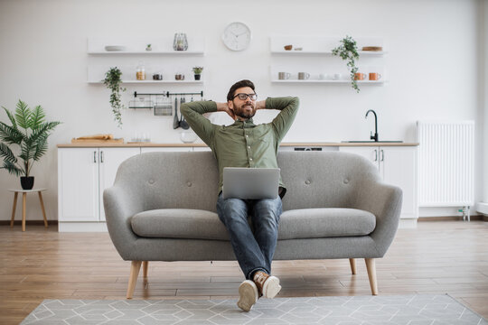 Positive man resting with hands behind head while leaning on cozy sofa and dreaming about vacation. Caucasian bearded male in eyeglasses and casual attire enjoying remote work while staying at home.