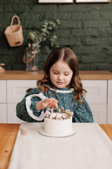 Adorable little child girl wears festive dress takes a bite out of a decorated flowers name cake at a birthday party. Happy smiling kid licks white cream from her dirty face and shows thumb up indoors