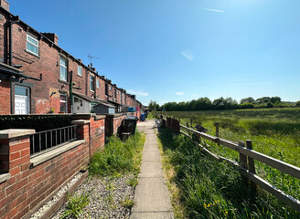Fototapeta na wymiar Pathway, close to a row of, old red brick terrace houses, overlooking a field, with wild plants and grasses in, Littleborough, UK