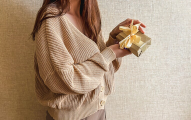 An unrecognizable brunette girl in a beige sweater holds a gift with a ribbon in her hands. a small gift in honor of the holiday