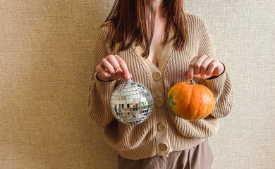 An unrecognizable brunette girl in a beige sweater holds a silver disco ball and an orange pumpkin in her hands. Halloween or Thanksgiving House Party