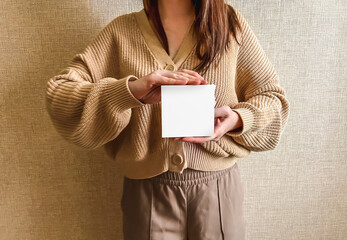 An unrecognizable brunette girl in a beige sweater holds a white square mockup in her hands. An empty sign in the hands of a girl