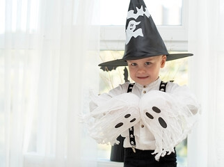 Halloween concept. A small handsome boy in a wizard's hat with white ghosts plays cheerfully and emotionally in a home interior against the background of a window.