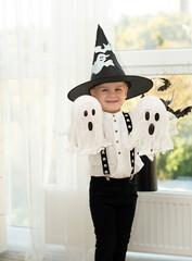 Halloween concept. A small handsome boy in a wizard's hat with white ghosts plays cheerfully and emotionally in a home interior against the background of a window.