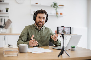 Popular blogger in wireless headphones talking to camera and using mobile on tripod at home. Caucasian smiling influencer making educational video vlog and taking notes at desk.