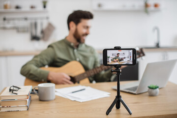 Fototapeta na wymiar Skillful musician sitting at home office using smartphone and tripod for filming video tutorial about guitar playing. Handsome male blogger sharing with followers in social media artistic experience.