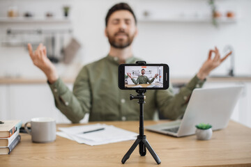 Blurred background of male streamer meditating with closed eyes and holding mudra gesture while filming online course at home. Focus on wireless smartphone fixed on tripod and recording video.