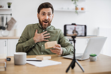 Emotional young guy opening cardboard box and looking inside with excitement while filming video on smartphone. Caucasian male blogger sharing positive emotions with subscribers in social networks.
