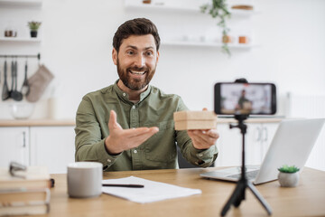 Emotional young guy opening cardboard box and looking inside with excitement while filming video on smartphone. Caucasian male blogger sharing positive emotions with subscribers in social networks.