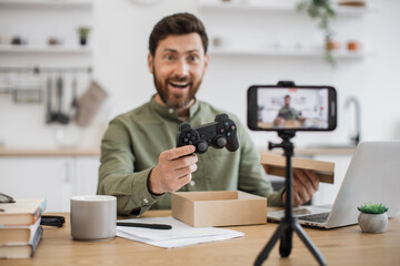 Thrilled male vlogger looking at mobile camera with wireless joystick in hand after opening cardboard box. Bearded man sharing sincere emotions with followers of parcel unpacking during live stream.