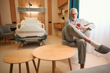 Man in hospital pajamas sits with mobile phone on soft chair