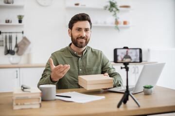 Caucasian young vlogger recording new video on smartphone about unboxing modern technologies. Bearded male person holding box and enjoying reviewing online purchase. Concept of working at home.