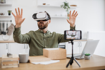 Excited caucaisan blogger testing VR headset while recording video on modern smartphone fixed on tripod. Bearded man making review of new gadget during unpacking of online purchase at home.