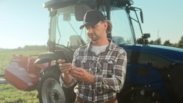 Tractor driver using smartphone. Farmer wearing checkerd shirt and cap. Man standing at field and texting. Harvest season. Male standing at background of tractor machine.