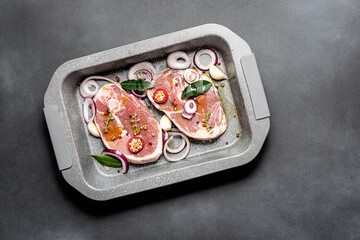 Raw organic lamb steaks with herbs, spices, garlic and onion rings in the oven pan on dark concrete table top view.