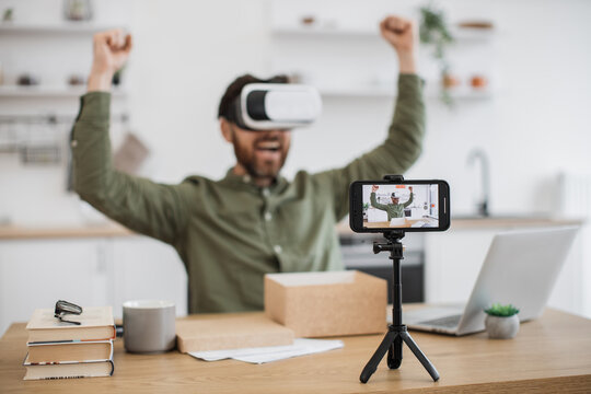 Modern smartphone recording video of happy male blogger testing new VR headset and making review for subscribers on social media. Innovative electronics and online purchase concept.