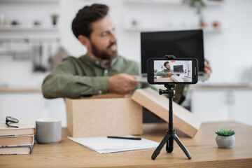 Modern smartphone fixed on tripod filming video of positive bearded man doing unpacking of new purchased laptop at home. Smiling caucasian vlogger giving feedback and advice about modern gadget.
