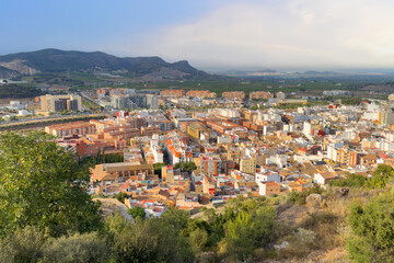 Fototapeta na wymiar View of the city of Sagunto in Spain. Buildings, houses and streets in city. View of rooftops and streets. Town against backdrop of mountains. Roofs of houses and roofs from side of Sagunto Castle.