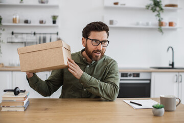 Excited caucasian man in casual wear and glasses sitting at home office with cardboard box in...