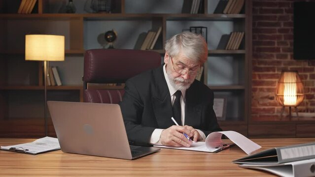 Confident business person in glasses and formal suit signing printed document at wooden desk in upscale office. Professional entrepreneur approving corporate agreement via signature in workplace.
