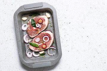 Raw organic lamb steaks with herbs, spices, garlic and onion rings in the oven pan on light concrete table top view.