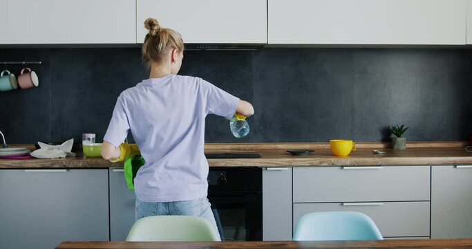 A young girl is cleaning the kitchen and dancing, turning and leaning on the table, taking a deep breath of fatigue.