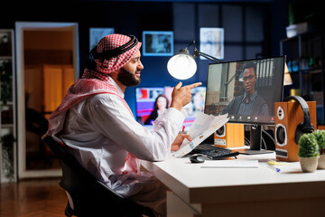 Picture of a Muslim man handling paperwork while chatting with his African-American coworker. Two young businesspeople are on a desktop computer doing a video call. - obrazy, fototapety, plakaty