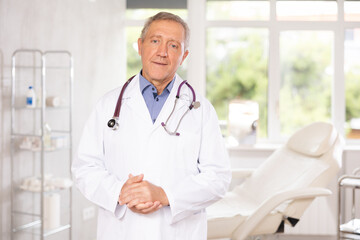 Portrait of smiling elderly man, confident professional doctor wearing white coat standing in modern light medical office..