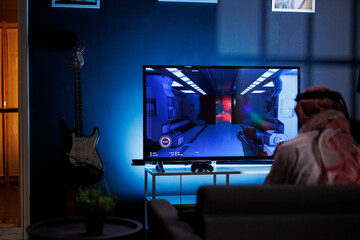 Rear-view shot of person playing with modern technology gadget using video games console on television. Muslim gamer with wireless controller sitting at home having fun with digital action.