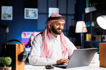 Young Arab man works diligently at his desk, browsing the internet on his digital laptop. Engrossed...