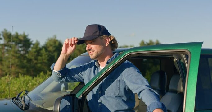 Handsome male farmer adjusting cap on head. Attractive farm worker leaning against pickup door. Man finished plowing or harvesting and enjoying sunset. Farmer standing at filed with truck.