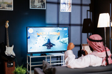 Middle Eastern man in traditional attire celebrates a victory, immersed in virtual reality game. With wireless controller, he grasps the joystick, capturing the excitement of online gaming.