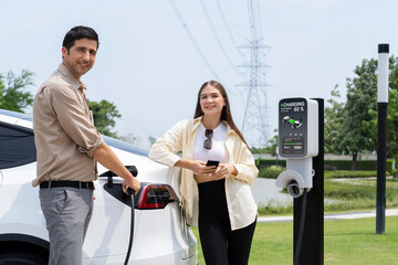 Young couple use smartphone to pay for electricity at public EV car charging station green city...