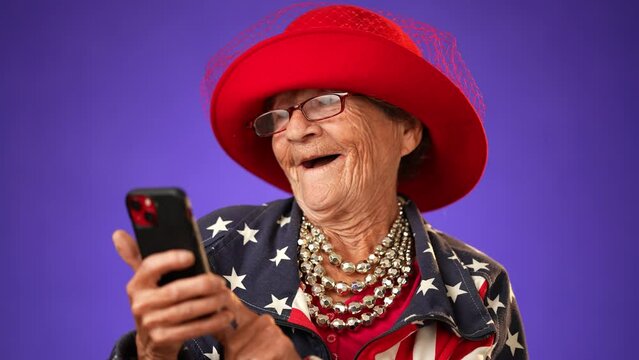 Closeup of funny crazy happy elderly old toothless woman shopping online using mobile cell phone wearing US flag jacket and glasses isolated on solid purple background studio portrait. 