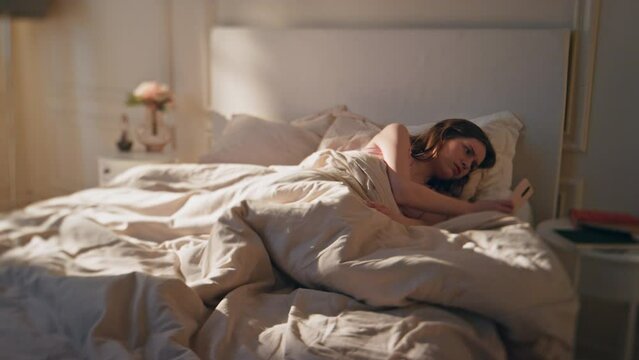 Sick woman lying home in bed morning. Tired awake female checking mobile phone