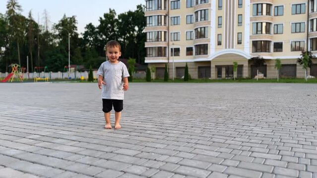 Beautiful Caucasian toddler boy walking outdoors barefoot. Kid looks at camera, talks and smiles.