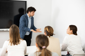 Naklejka na ściany i meble Male business coach standing near interactive board explaining topic while standing against group of people sitting at desks in front of him