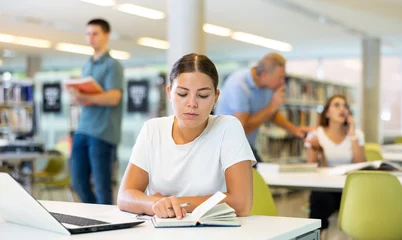 Fotobehang Concentrated young woman checking planner while studying in the library and using laptop © JackF