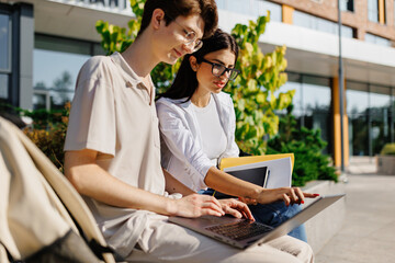 Happy young man and woman students sitting on park wall with laptop