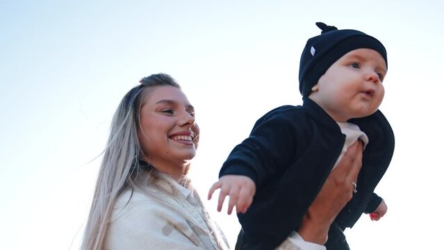 Happy smiling blonde Caucasian woman holding a baby in hands. Mother with her infant child outdoors. Low angle view.
