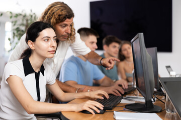 Interested young girl attending computer class, learning digital technology under guidance of positive adult male teacher standing nearby..
