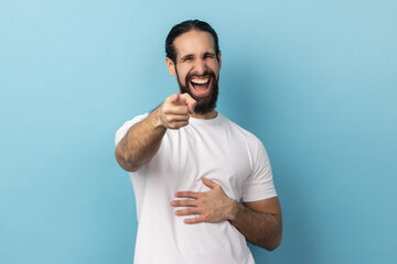 Well you're a joker. Portrait of positive handsome man with beard wearing white T-shirt pointing index finger at camera and laughing positively. Indoor studio shot isolated on blue background. - obrazy, fototapety, plakaty