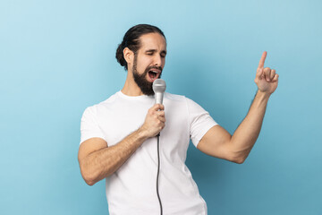 Portrait of funny man with beard wearing white T-shirt loudly singing song holding microphone in hand, having fun resting in karaoke, singer performance. Indoor studio shot isolated on blue background - obrazy, fototapety, plakaty