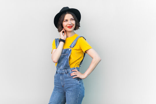 Portrait of smiling young adult hipster woman in blue denim overalls, yellow T-shirt and black hat, looking away, dreaming for something pleasant. Indoor studio shot isolated on gray background.
