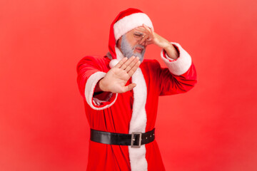 Santa claus in red costume pinching nose with fingers to avoid bad smell and showing stop gesture, demonstrating repulsion to stink, awful odor. Indoor studio shot isolated on red background. - obrazy, fototapety, plakaty