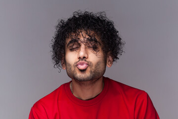 Fototapeta na wymiar Portrait of romantic flirting handsome bearded man with curly hair, standing with closed eyes, keeps lips pout, sending kissing. wearing red jumper. Indoor studio shot isolated on gray background.