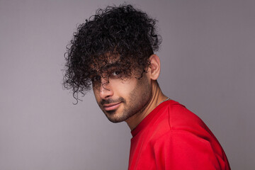 Fototapeta na wymiar Portrait of attractive handsome young adult bearded man with curly hair, wearing red jumper, looking at camera, showing his hairdo. Indoor studio shot isolated on gray background.